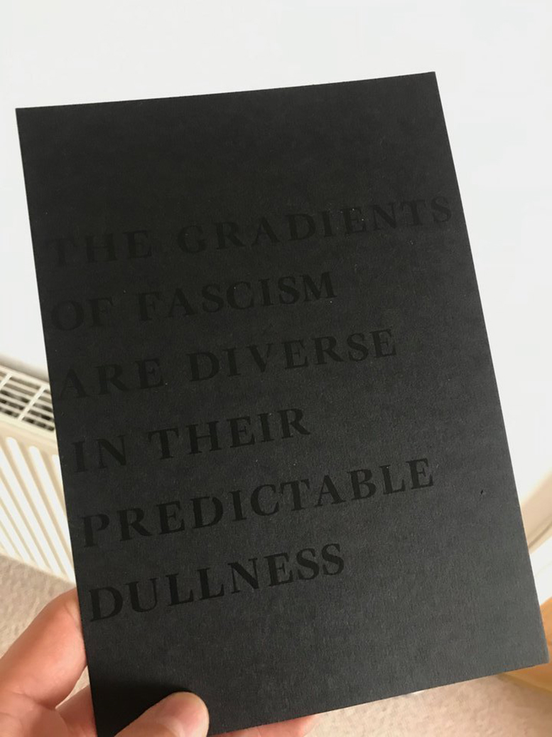 Hand holding black postcard with black text reading “THE GRADIENTS OF FASCISM ARE DIVERSE IN THEIR PREDICTABLE DULLNESS” image