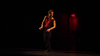 A woman wearing a red shirt and black pants dances on a dark stage.