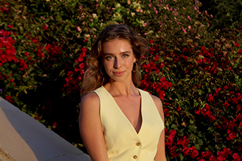 A young woman with a cream-colored top poses in front of a backdrop of leaves and flowers