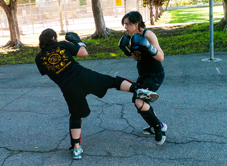 Two women wear boxing gloves and are engaged in a sparring contest on an asphalt lot, with one woman kicking the other.