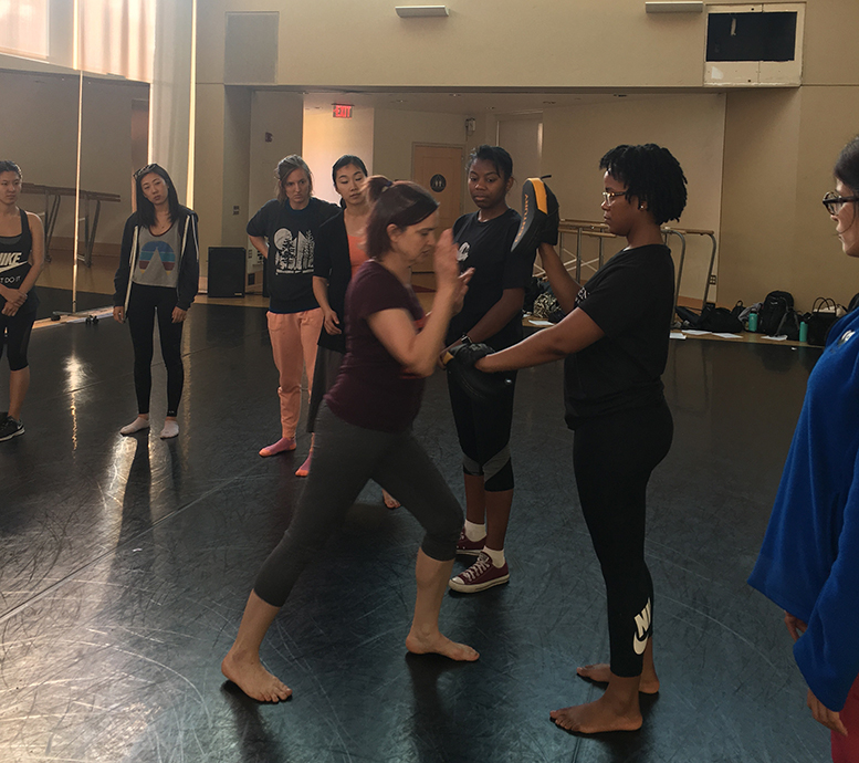 Two women engaged in a sparring contest, with Janet O'Shea preparing to kick the other woman's upheld glove, while several students look on.