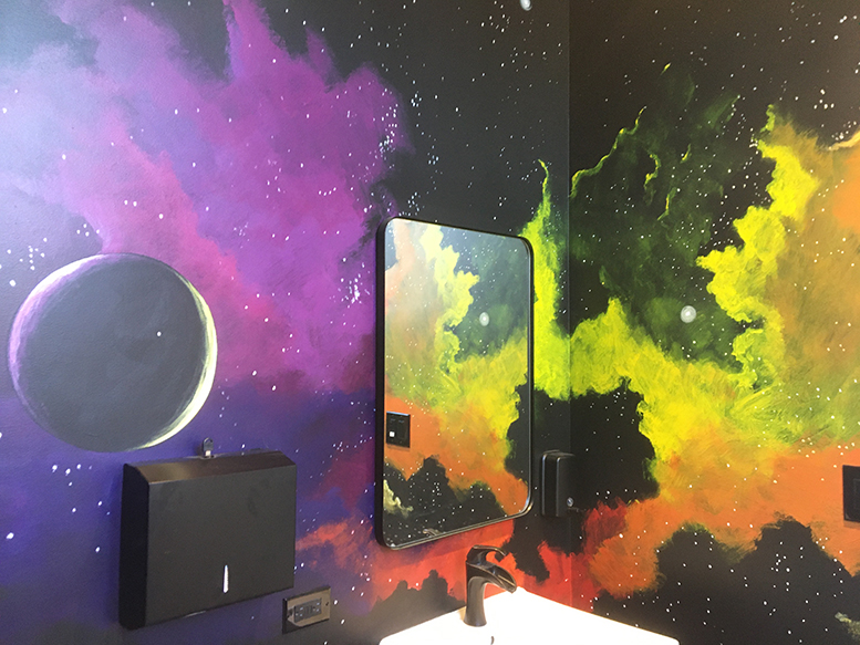 The restroom walls show vibrantly-colored planets and galaxies, with a mirror, sink and paper towel holder.