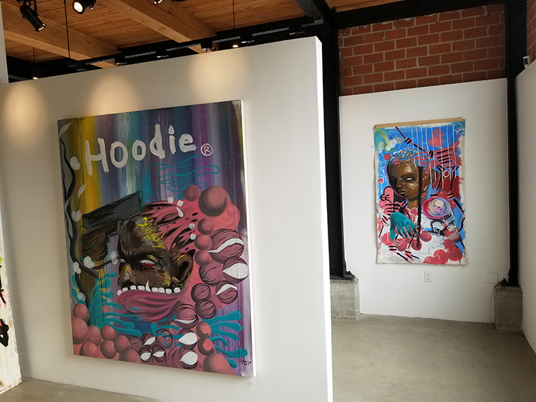 Image shows two paintings by Gianni Lee on view in Gallery 90220. One in the foreground shows a Black man with a gun to his head. Another in the background shows a Black woman.