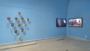 An image of Lili Flores Raygoza's exhibit "Behind the Mask." On blue walls hang a triangular display of painted masks to the left and two TV screens to the right. The screens display masks and images of dancers.