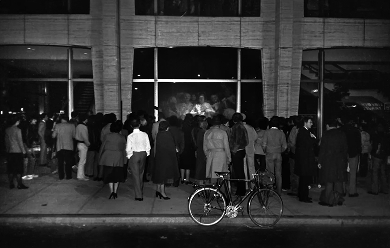 People assembled on the sidewalk stare at a video screen showing another group of people looking back at them.