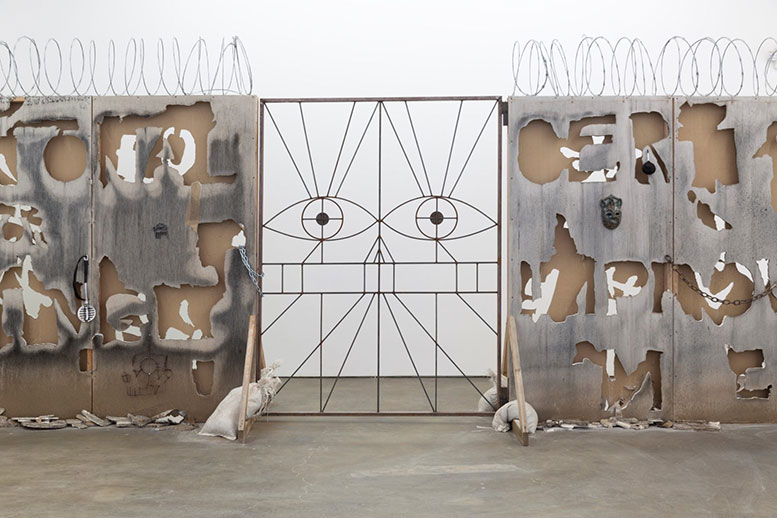 An installation that shows a metal gate with a face, and walls covered with barbed wire