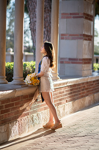 Yaying stands inside the entry of Royce Hall, facing away from the camera and looking out onto Royce Quad. She wears a white dress and a blue graduation sash.