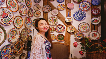 Yaying is shown torso-up standing in front of a wall in a rustic-looking building. The wall is covered with beautiful porcelain plates featuring floral designs in many colors.