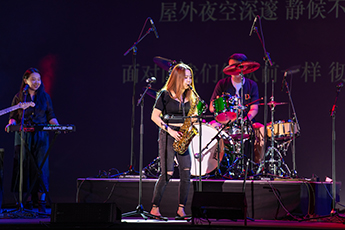 Yaying plays the saxophone, standing on a darkened stage. Behind her is a drummer and to her left is a smiling keyboardist.