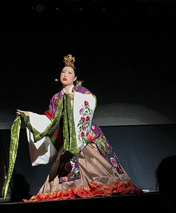 Kayla Tange pictured during a performance. Her arms are lifted, holding a piece of long green fabric as she glances towards stage left. She wears a colorful Korean hanbok with floral designs. Her cheeks are highlighted with bright red circles of blush and she wears a gold headpiece.