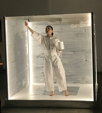 Kayla Tange during a performance. She wears loose-fitting, all white clothes, and is encased in a glass box with all sides but one painted a grey-white color. She holds a white paint can and paintbrush, and is painting the final glass side of the box with more white paint.