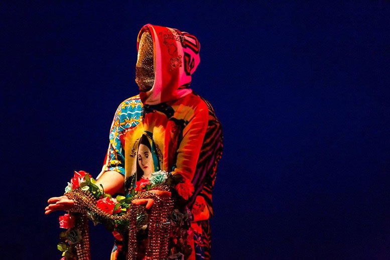 A photograph of a figure dressed in a hooded gown with the Virgin Mary on the front, and a face shield obscuring the face, and holding flowers and beaded strings