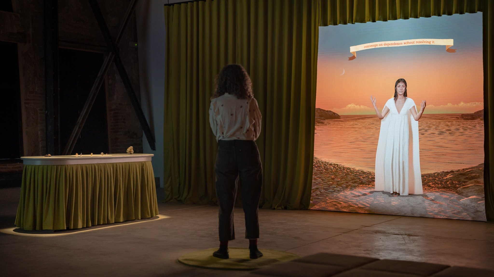 A person views an installation featuring a large digital screen displaying a woman in a white dress against a sunset beach scene.