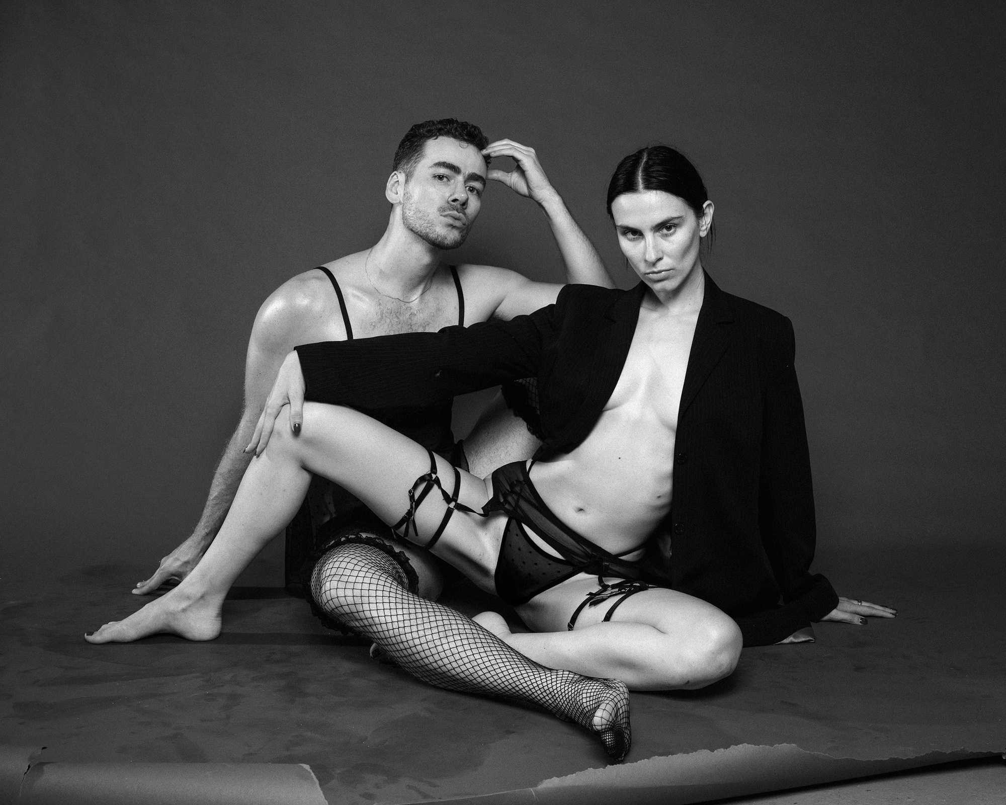 Black-and-white studio portrait of two seated people wearing black fashion garments and hosiery, posed against a plain dark backdrop.