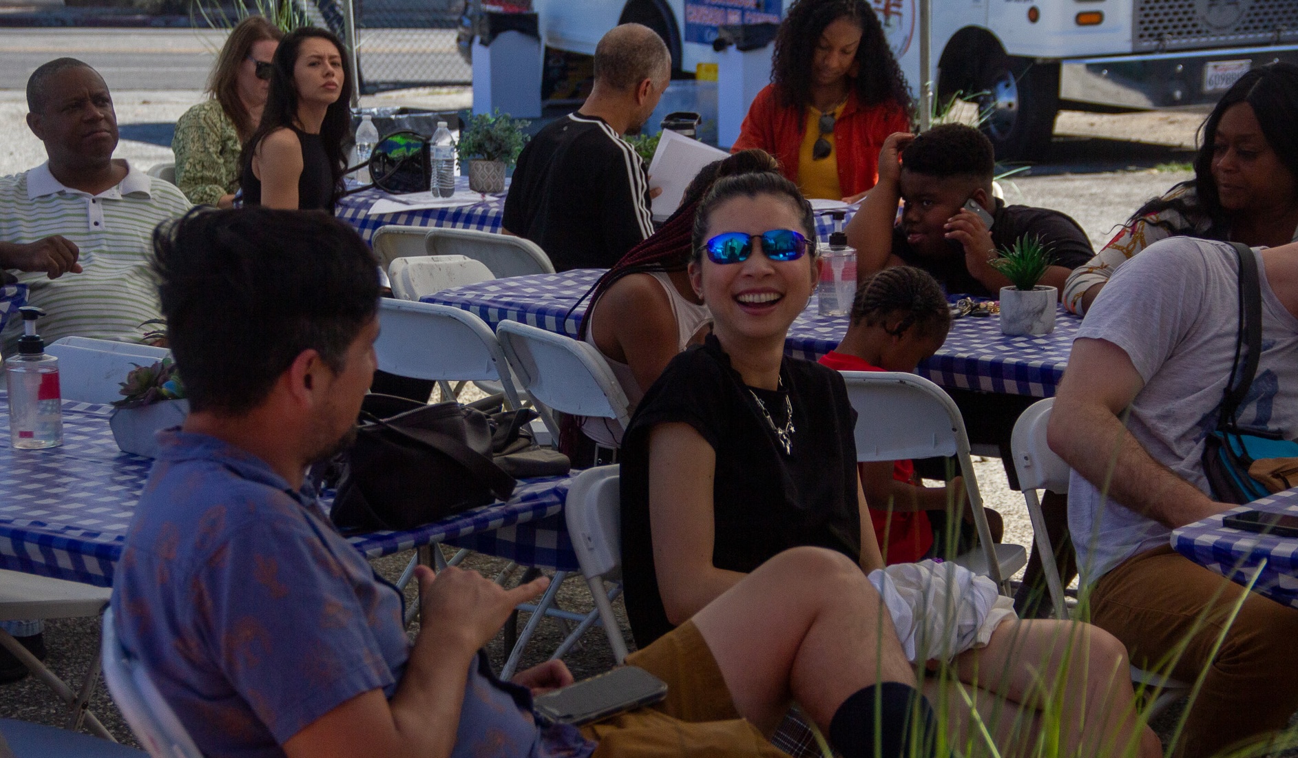 A group of people sits around outdoor tables with blue-and-white checkered tablecloths, chatting and eating.
