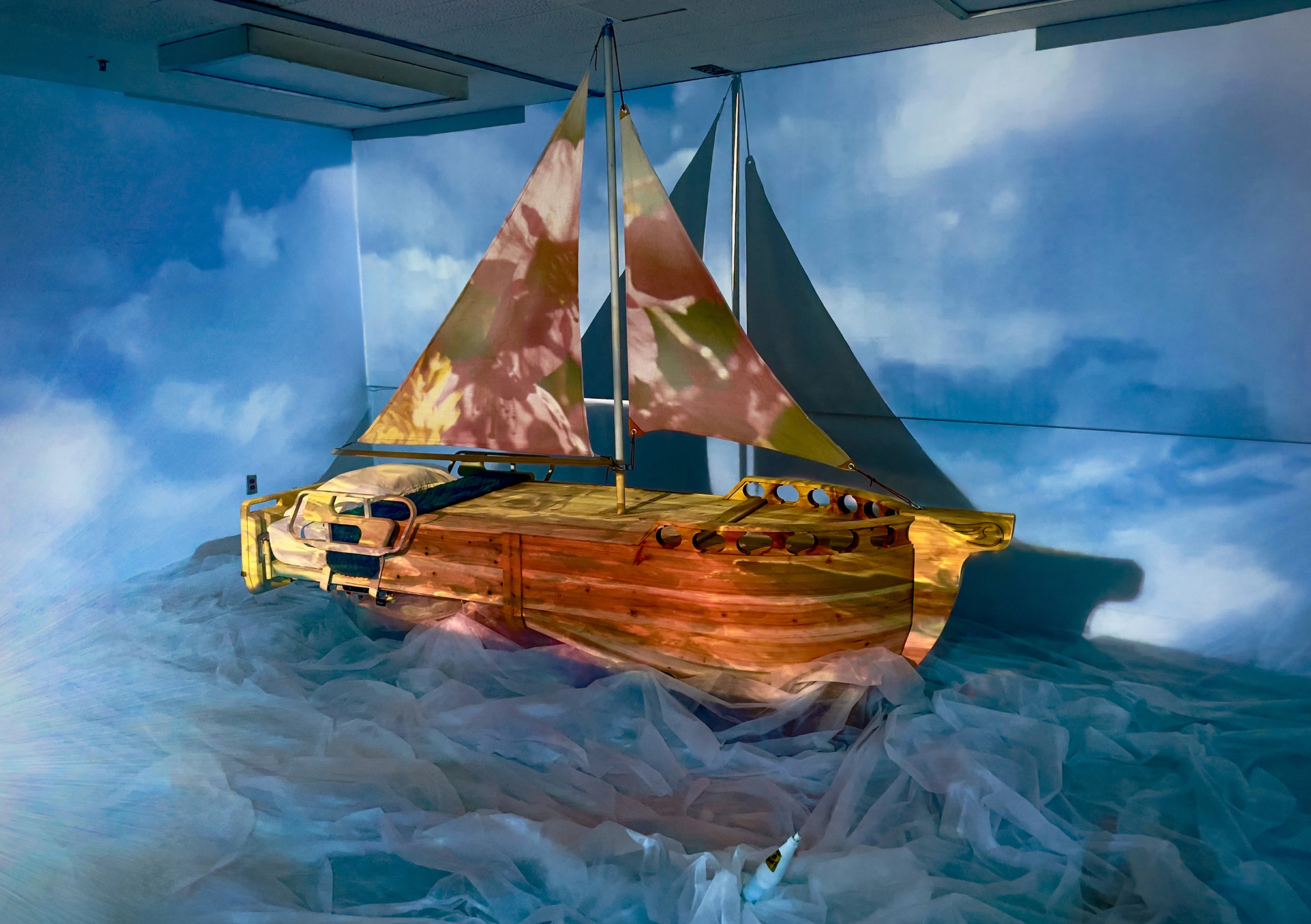 Wooden sailboat installation with projected sky and floral imagery, displayed indoors on a floor covered with translucent fabric resembling water.