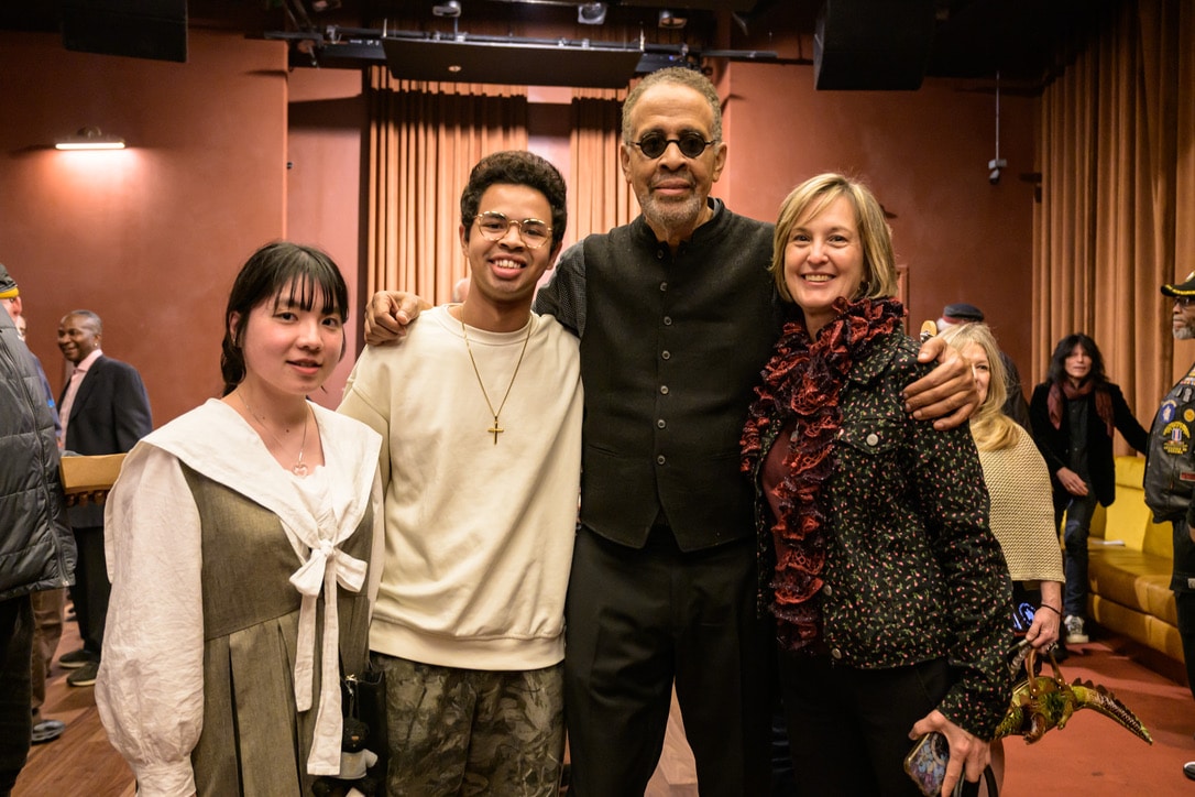 Four people pose together at an indoor event venue, standing in front of rows of seating and stage curtains, with other attendees visible in the background.
