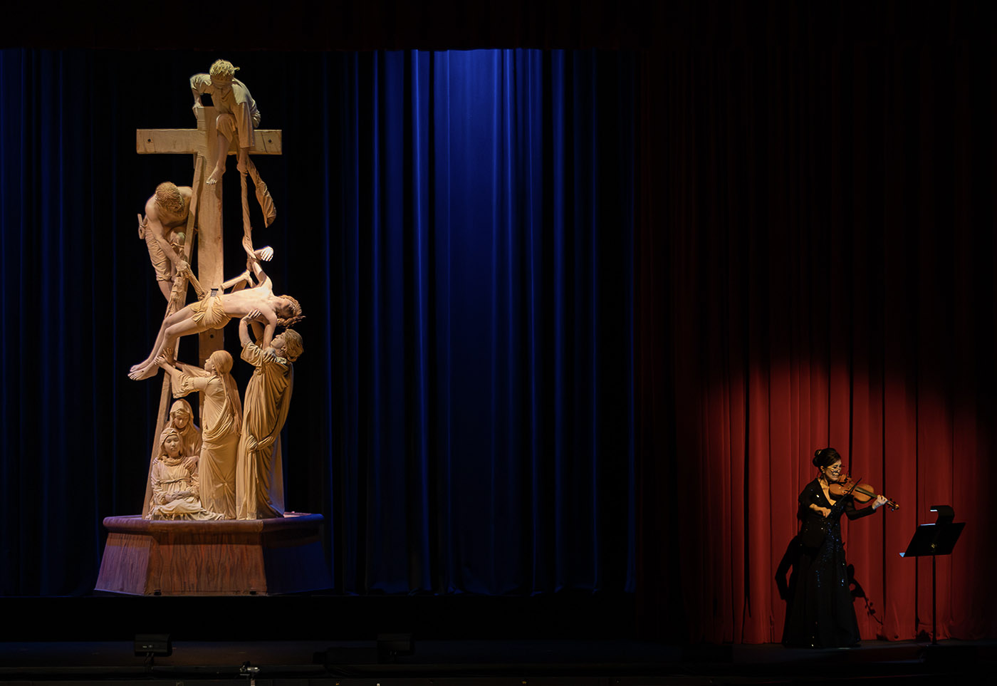 A dramatic stage scene: an ornate crucifixion sculpture stands beneath blue lights while a violinist performs against red curtains.