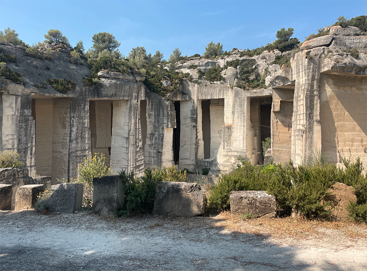 Sunlit ancient limestone quarry in France with carved doorways and stone chambers, weathered rock walls, shrubs, and trees beneath a clear blue sky.