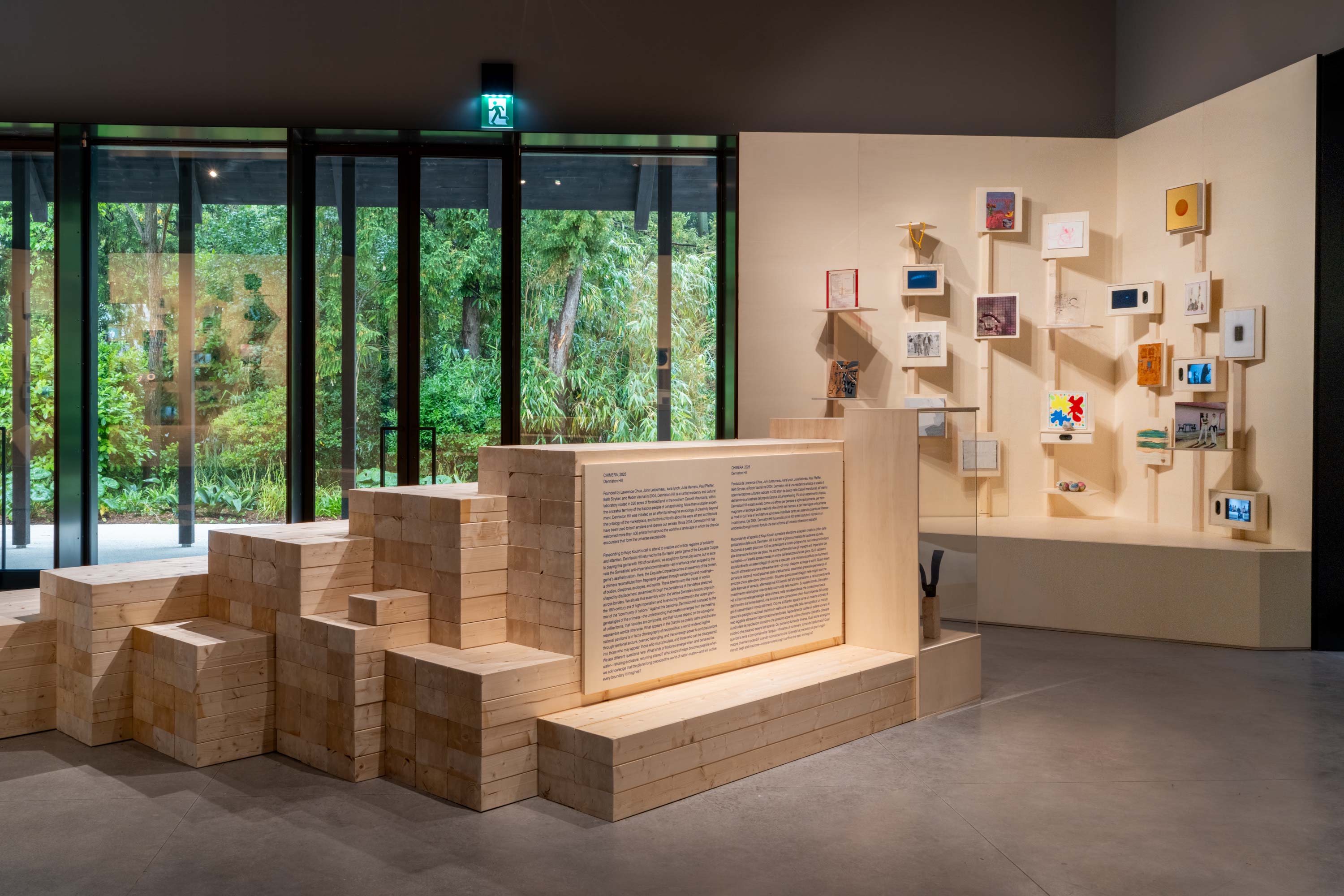 Museum gallery with a stepped wooden installation and display wall featuring small artworks, set beside floor-to-ceiling windows overlooking a lush garden.