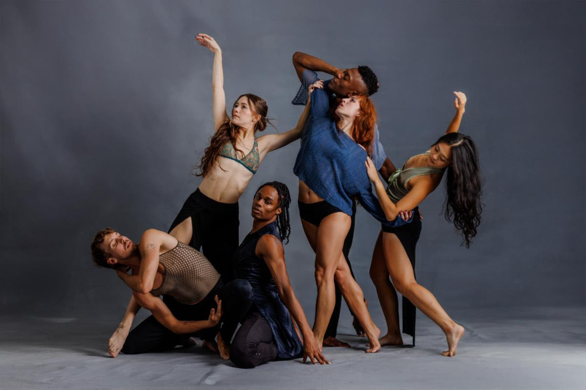Five contemporary dancers pose together in an expressive group formation against a gray studio backdrop.