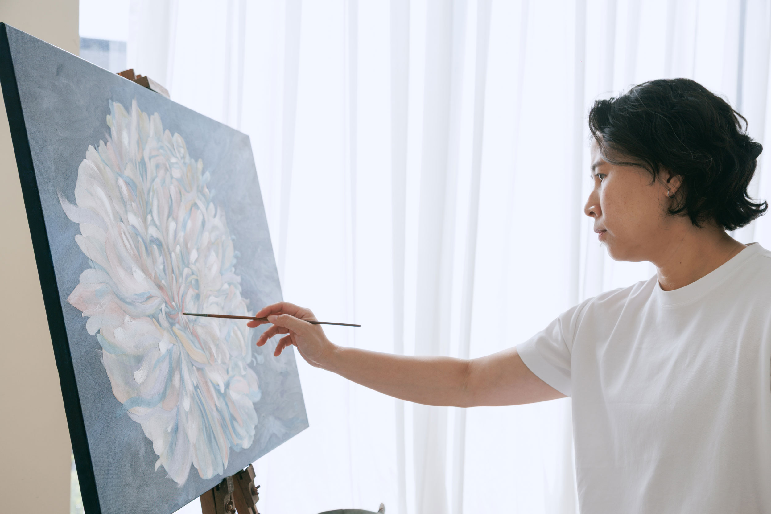 Person painting a large floral artwork on an easel in a bright studio with soft natural light.