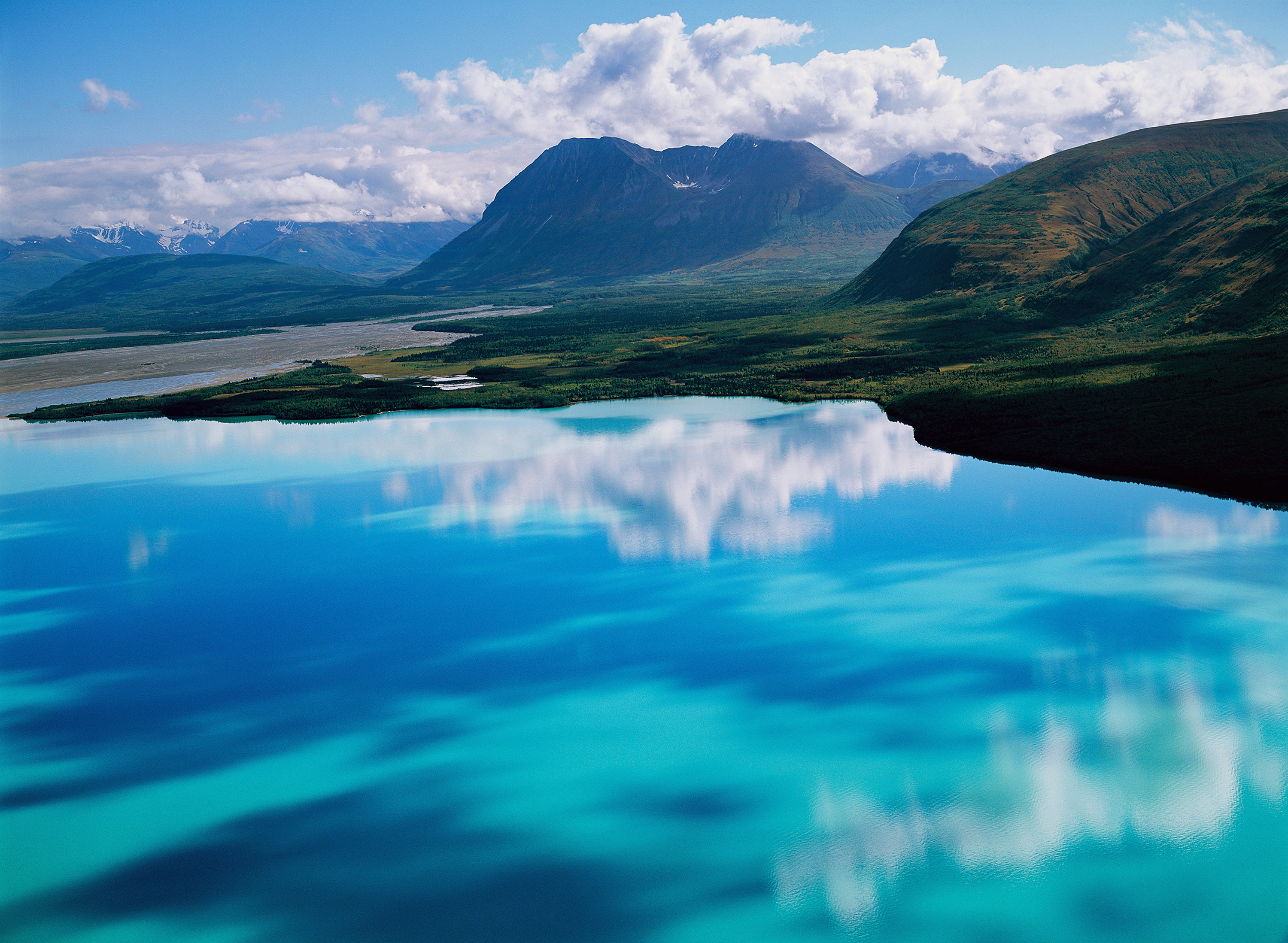 Aerial view of a turquoise lake reflecting clouds and surrounding mountains beneath a bright blue sky.