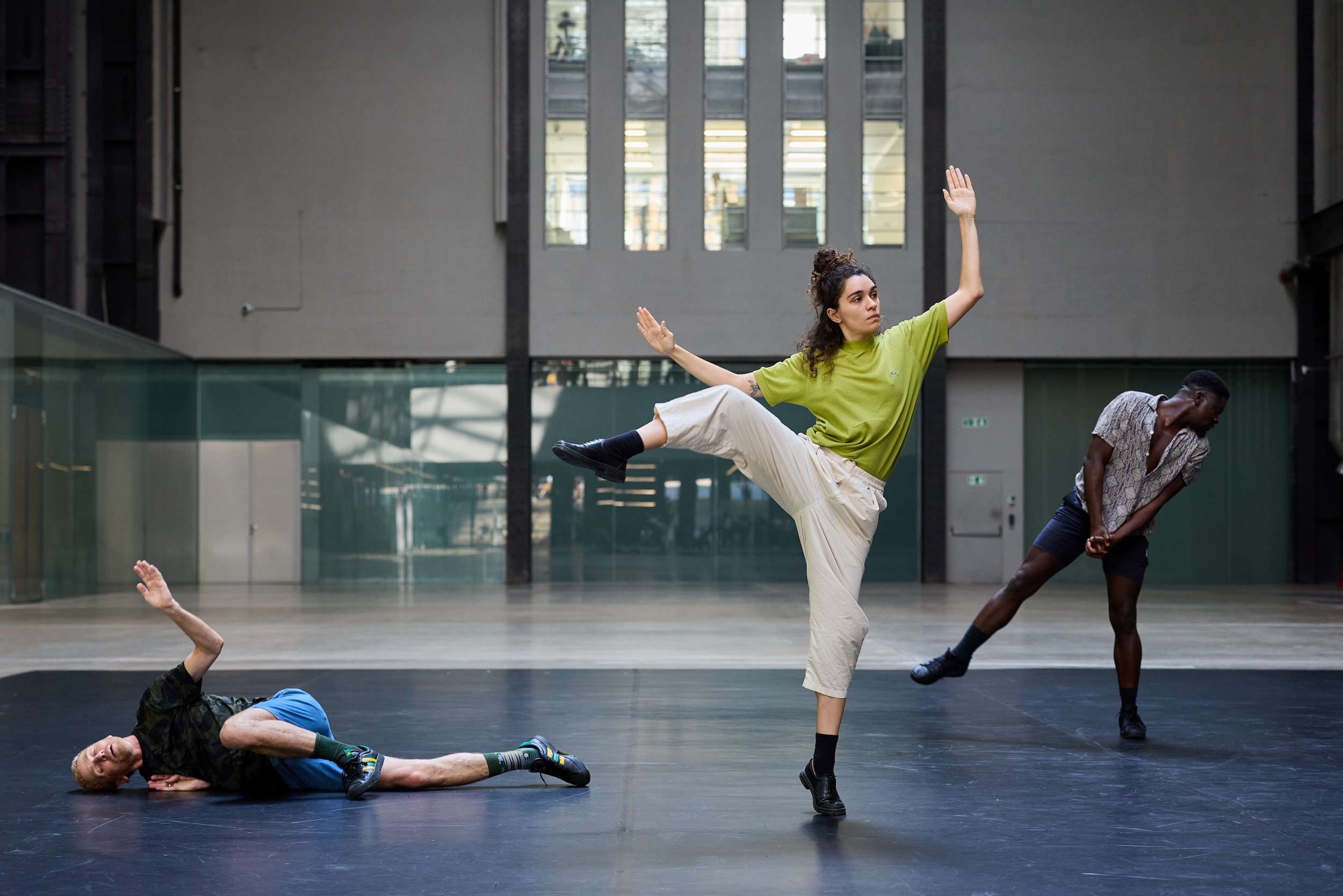 Three dancers hold diverse poses in a large dance rehearsal room.