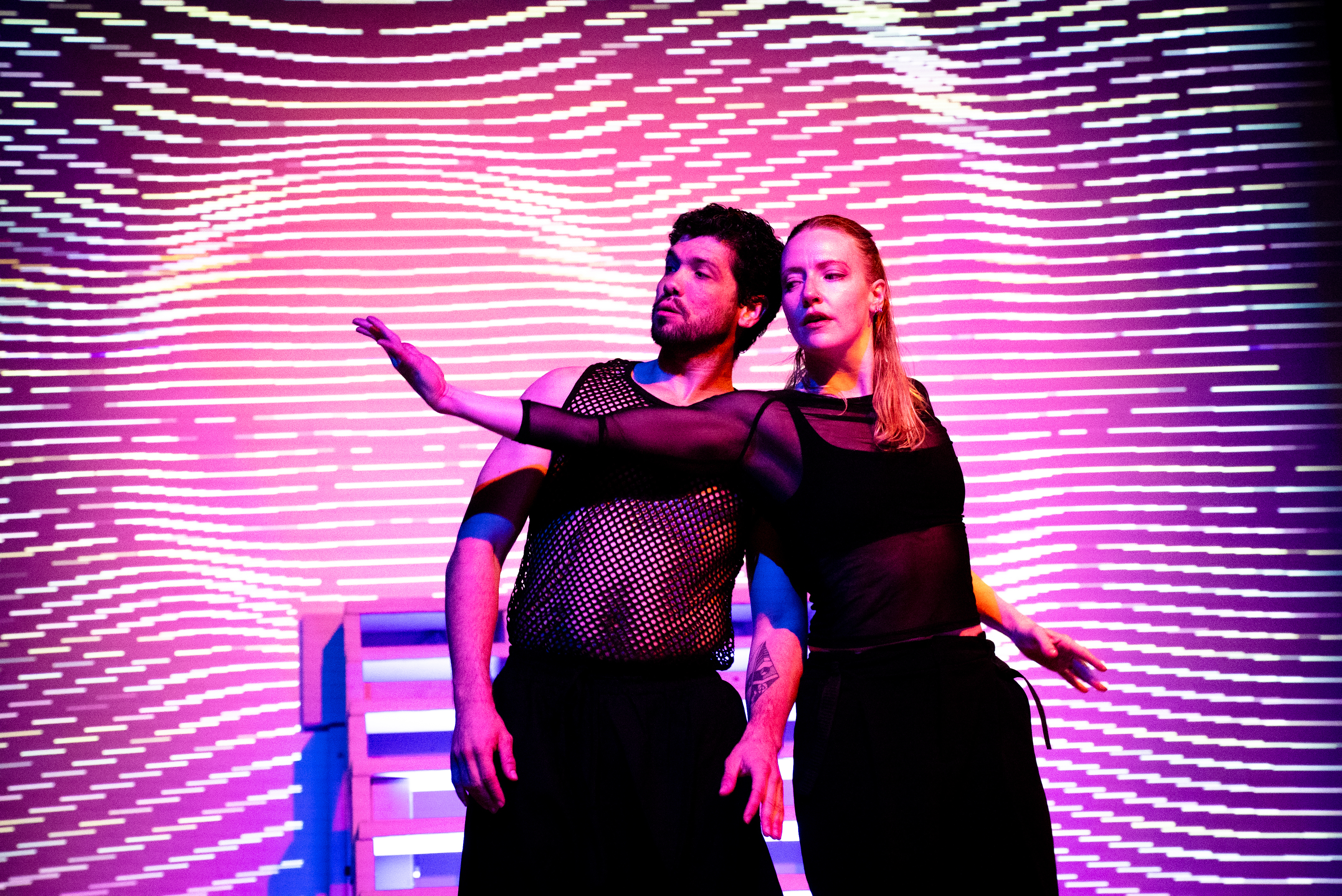 Two performers pose in front of a vibrant pink and purple digital projection with wavy light patterns.