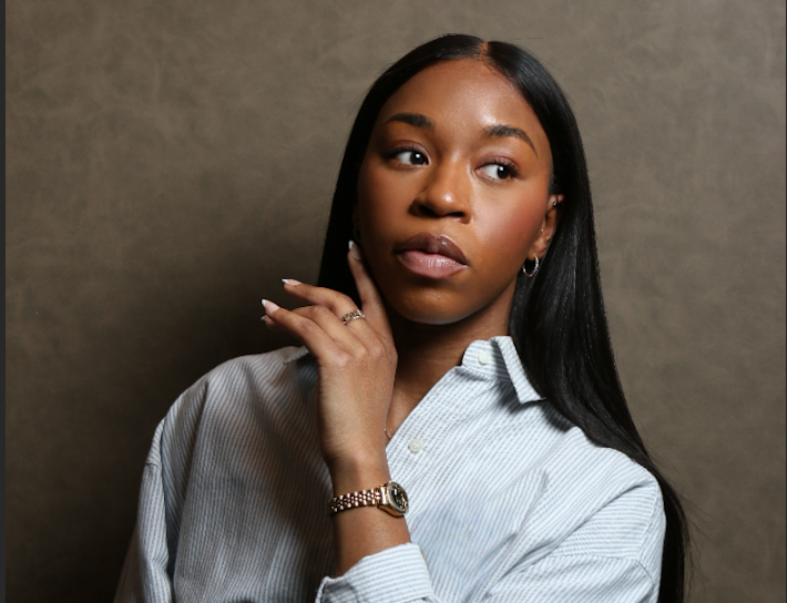 Studio portrait of a person with long hair wearing a striped button-down shirt, posed against a neutral textured backdrop.
