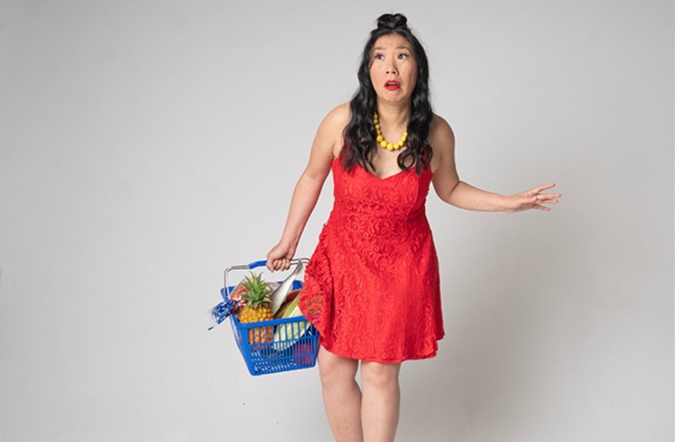 Person in a red dress carrying a grocery basket while assorted fruits and vegetables are scattered across the floor in a studio setting.
